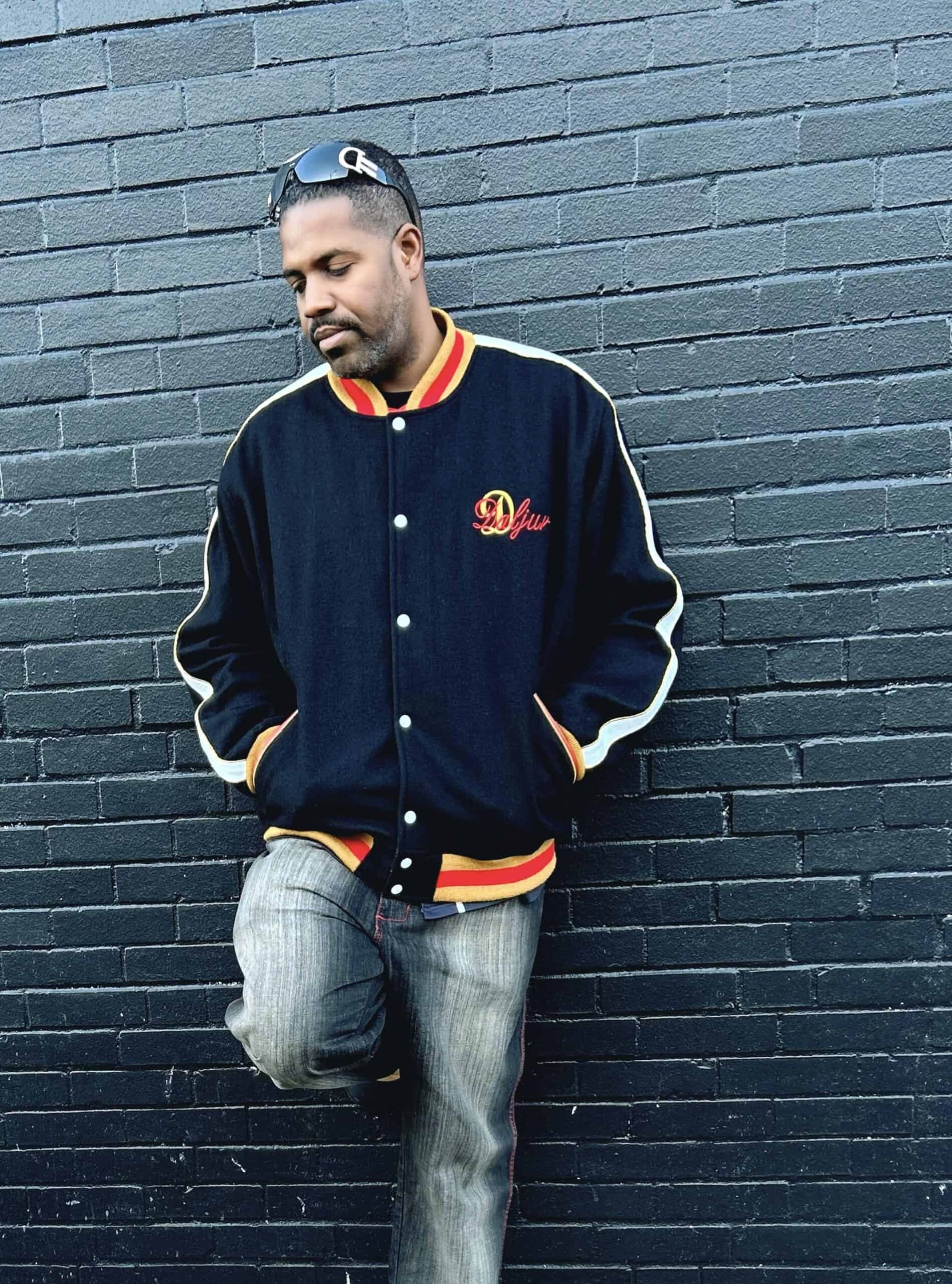 A man standing against a brick wall wearing jeans and an orange jacket.